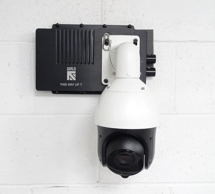 Une grande caméra de surveillance blanche et noire fixée sur un mur, à côté d'un dispositif indiquant 'Made in Britain' et 'This Way Up ↑'.