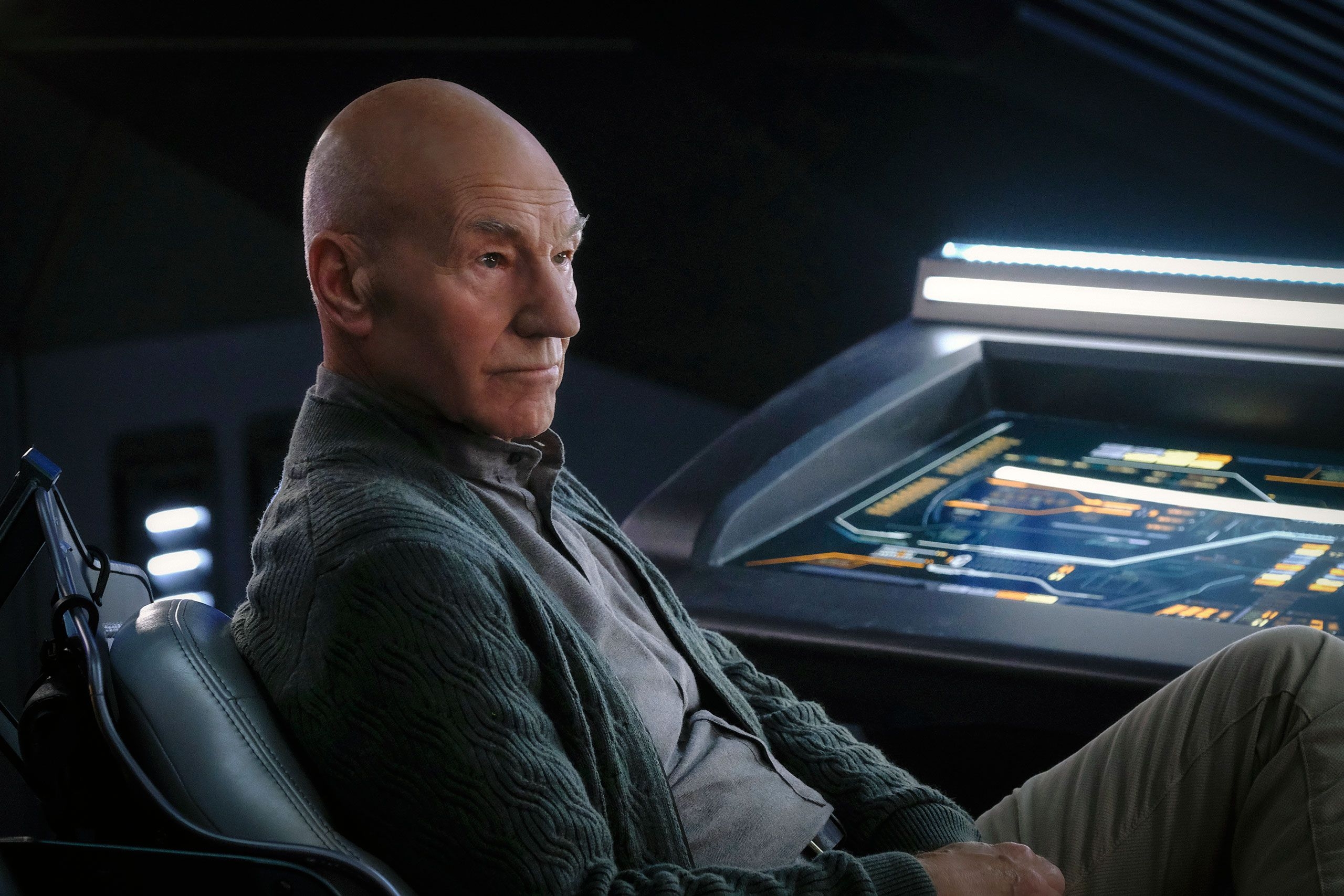 A person sitting in a futuristic room with a digital control panel displaying various orange and white interface elements on a screen.